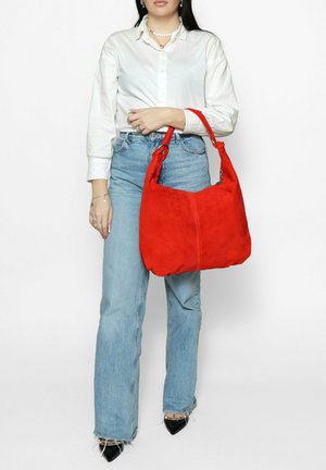 Rode suède hobo-tas met een gebogen bovenkant, zachte textuur en een enkele schouderriem, vastgehouden door een persoon die een wit overhemd en blauwe jeans draagt.