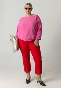 Roze top met blote schouders, rode op maat gemaakte broek, zwarte puntige schoenen, en een metalen zilveren tas in de hand. Eenvoudige accessoires en strakke lijnen komen naar voren.