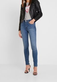 Schwarze Leder-Bikerjacke, graues T-Shirt mit rotem Logo, blaue schmal geschnittene Jeans und schwarze Sandalen mit Riemchen und Absatz. Minimalistisches Design.