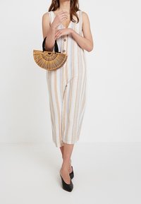 Striped, sleeveless linen dress with buttons, featuring vertical beige and white stripes. Holding a bamboo half-moon bag with a black handle.