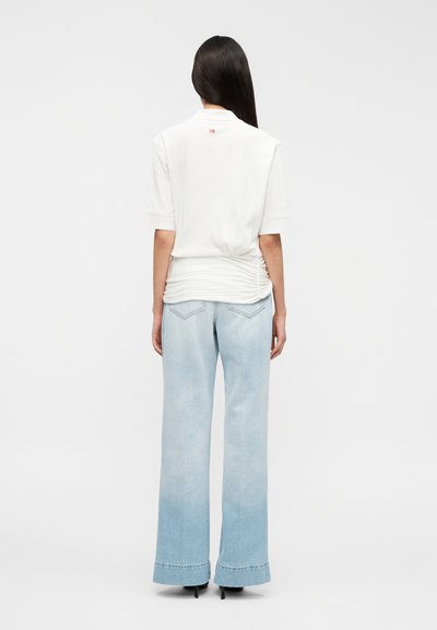 Haut blanc à manches courtes avec un détail froncé à l'arrière et un logo rouge. Associé à un jean en denim bleu clair à jambes larges avec une finition délavée.
