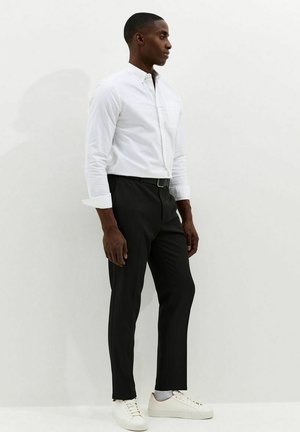 Camisa blanca abotonada con bolsillo en el pecho, combinada con pantalones negros y zapatillas blancas. La camisa tiene las mangas remangadas y un corte entallado.