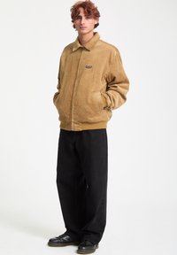 Veste en velours côtelé beige avec un zip à l'avant, un col pointu et des poignets côtelés. Pantalons noirs et chaussures noires complètent la tenue. Design simple, coupe décontractée.
