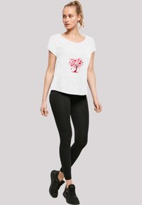 Weißes T-Shirt mit kurzen Ärmeln und pinkem Baumdesign, kombiniert mit schwarzen Leggings und schwarzen Sneakers, vor einem neutralen Hintergrund.