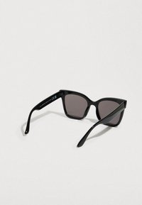 Μαύρα γυαλιά ηλίου με γυαλιστερό φινίρισμα, γωνιώδη σχήμα cat-eye, σκούρους γκρι φακούς και λεπτά μπράτσα. Διαθέτουν διακριτικό λογότυπο στο πλαϊνό μέρος.