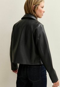 Vrouw gezien van achteren in een zwart leren jasje en donkerblauwe jeans tegen een effen lichte muur als achtergrond.