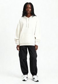 Noor mustanahast naine palmikutega juustega, kandes liibuva kreemja kapuutsi, musti tänavapükse ja valgeid tossusid, seisab näoga ettepoole.