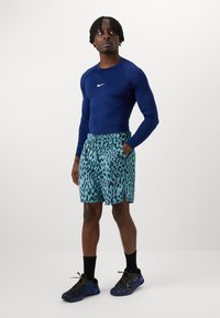 Tamsiai mėlynas ilgomis rankovėmis sportinis marškinėliai su balta logotipu; turkio spalvos šortai su juodu leopardų raštu; juodi sportiniai batai su geltonais akcentais.