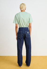 Ljusgrön bomullströja med avslappnad passform, ihopparad med mörkblå denimbyxor som har två bakfickor och vita sömmar.