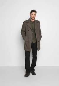 Abrigo de cuadros marrón de doble botonadura con grandes solapas, llevado sobre una camisa verde oscura, combinado con pantalones negros y zapatos oscuros, sobre un fondo neutro.