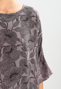 Blusa floreale in morbido tessuto grigio. Presenta un colletto rotondo, maniche corte e un motivo stilizzato di papaveri neri su uno sfondo testurizzato.