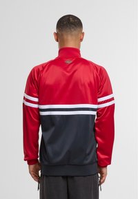 Rød og marineblå løbejakke med høj krave, hvide vandrette striber og ribbede manchetter. Har lynlåslommer og et struktureret logo-accent.