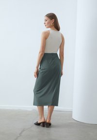 Sleeveless beige top paired with a high-waisted, floor-length dark green skirt. Material is smooth with subtle draping; block-heeled black mules.