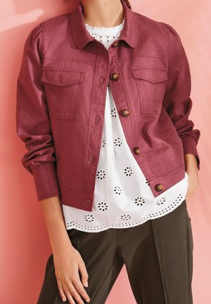 Une personne porte une veste bordeaux boutonnée par-dessus un chemisier blanc à œillets avec des découpes florales et un pantalon foncé, sur un fond rose.