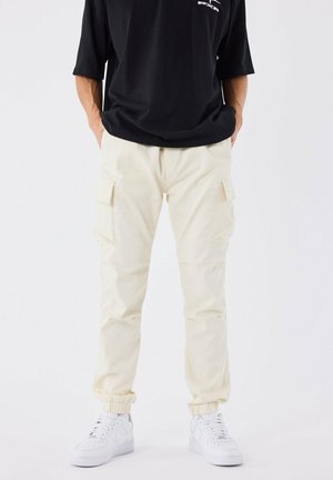 Pantaloni cargo beige con polsini elastici e tasche laterali, abbinati a una t-shirt nera oversize e sneakers bianche. Tessuto della stoffa morbido.