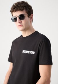 Černé bavlněné tričko s bílým obdélníkovým grafickým motivem s nápisem "Calvin Klein NEW YORK" na levé straně hrudi. Model nosí černé sluneční brýle.