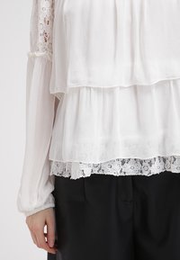 Blouse blanche à couches avec tissu transparent, bordures en dentelle et manches longues. Le design comprend une silhouette fluide et des détails délicats.