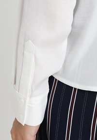 Witte blouse met lange mouwen en knoopmanchetten, met een gladde textuur. Gecombineerd met marineblauwe stof met verticale strepen en rode accenten.