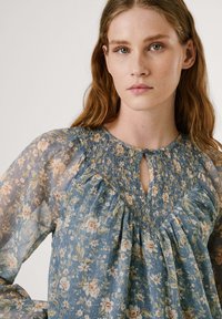 Blusa azzurra fluida con motivo floreale, scollatura arricciata e chiusura a bottone nella parte centrale. Maniche lunghe trasparenti con una delicata texture.