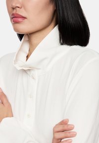 Weißes Hemd mit hohem, gefaltetem Kragen, ausgestattet mit einer Knopfschließe und einer glatten, weichen Textur. Die Ärmel sind lang und locker.