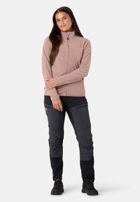 Jasnoróżowa polarowa kurtka z suwakiem z przodu, noszona na ciemnych, dwutonowych spodniach cargo z wieloma kieszeniami i naszywką z logo. Czarne obuwie.