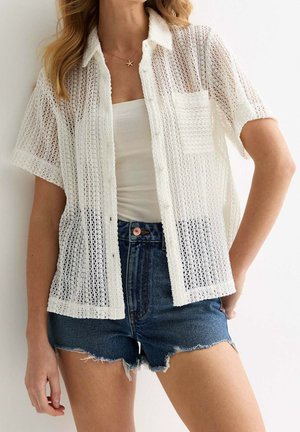 Weißes, kurzärmeliges Hemd mit offenen Maschen und Knöpfen, mit Tasche, getragen über einem weißen Tanktop, kombiniert mit dunklen Jeansshorts.