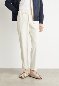 Pantalones beige claro con cintura elástica y cordón, combinados con sandalias beige con tiras de acento. Chaqueta azul marino parcialmente visible.