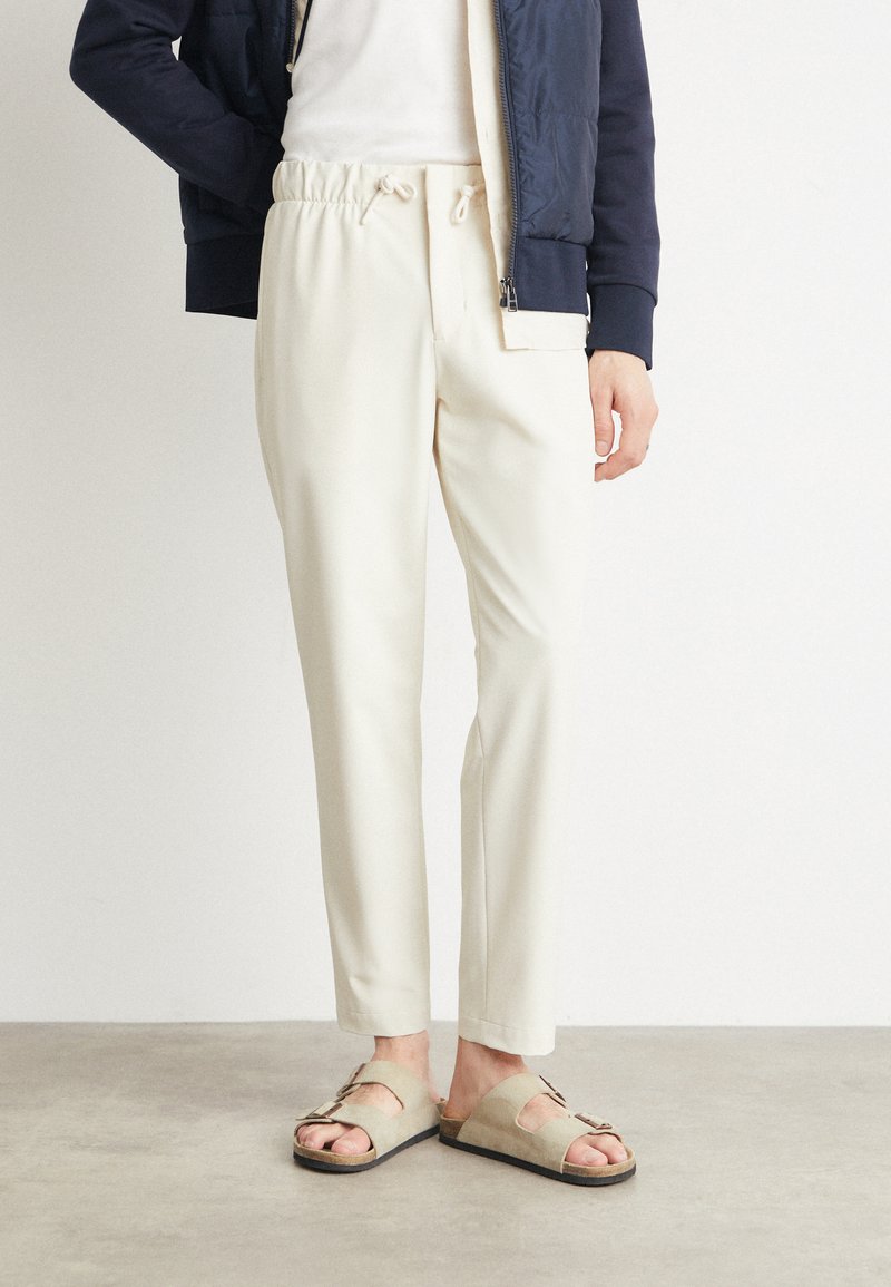 Pantalones beige claro con cintura elástica y cordón, combinados con sandalias beige con tiras de acento. Chaqueta azul marino parcialmente visible.