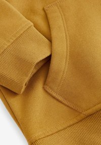 Sweatshirt jaune moutarde avec des poignets côtelés et un détail de poche. Texture douce avec des accents de couture visibles pour la durabilité.