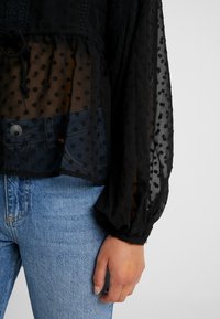 Personne portant un jean bleu clair et un chemisier noir transparent à manches longues avec un motif à pois, la main détendue le long du corps.