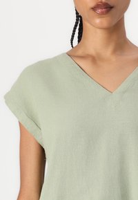 Top a maniche corte e scollo a V in morbido tessuto verde chiaro. Presenta una vestibilità comoda con cuciture in evidenza lungo lo scollo e le maniche.