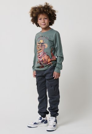 Groene longsleeve shirt met een afbeelding van een cowboydino, gecombineerd met een donkere cargobroek en witte sneakers met blauwe accenten.