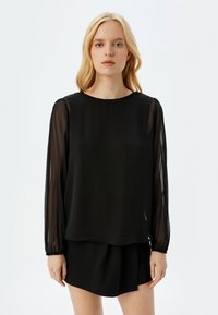 Top negro de manga larga con mangas de tejido transparente, escote redondo y corte holgado. Textura suave; combinado con una falda negra.