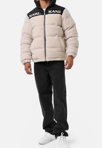 Giacca puffer in fleece beige chiaro con pannello nero sulle spalle che presenta la scritta "KARL KANI". Abbinata a jeans neri e sneaker bianche.