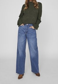 Breda blå denimjeans med hög midja och fem fickor, tillsammans med en mörkgrön stickad tröja. Bruna klackboots.