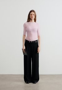 Γυναίκα που στέκεται μπροστά σε λευκό τοίχο, φοράει ροζ ριμπ πλεκτό τοπ, φαρδιά μαύρη παντελόνα με ζώνη, κρατώντας μικρό μαύρο τσαντάκι στο αριστερό χέρι.