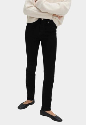 Osoba v černých úzkých džínách, krémovém svetru a černých plochých botách stojící se založenýma rukama na jednobarevném pozadí.