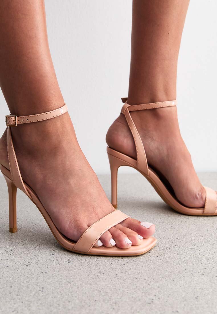 Nude hoge hakken sandalen met enkelbandjes, open teen ontwerp, gladde lakafwerking en slanke stiletto hakken, geplaatst op een lichte ondergrond.