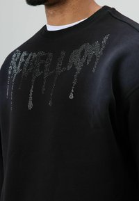 Zwarte sweatshirt met een ronde hals en een zilverkleurige strass-steengrafiek met een druppelend ontwerp in het midden. Zachte stofafwerking.