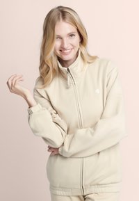 Beige zip-up jack met een zachte stof, hoge kraag, elastische manchetten en een logo op de borst, met een ontspannende pasvorm en minimale ontwerpdetails.