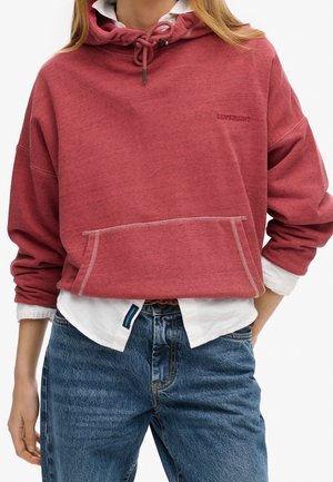 Woman wearing a red cropped hoodie with drawstrings over a white shirt and blue jeans, standing with one hand relaxed by her side.