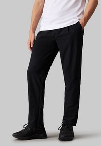 Pantaloni sportivi neri con superficie textured, caratterizzati da una vestibilità comoda, tasche laterali e una vita elastica, abbinati a sneakers nere.