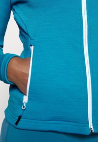 Main dans la poche d'une veste zippée couleur sarcelle avec des détails de fermeture éclair blancs, montrant un gros plan de la texture du tissu et des coutures.