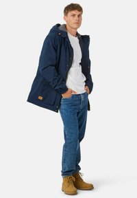 Marineblauwe waterdichte jas met een grijze gevoerde capuchon, gecombineerd met een witte blouse en blauwe denim jeans. Bruine veterschoenen maken de look af.