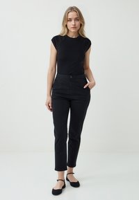 Une femme se tient contre un mur clair, portant un haut noir ajusté et un pantalon noir taille haute, avec une expression neutre et les mains dans les poches.