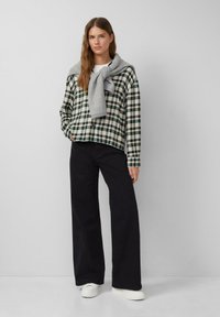 Karierte Button-Down-Bluse in Grün und Creme, kombiniert mit schwarzen, weiten Hosen. Grauer Strickpullover, lässig über die Schultern gelegt. Weiße Schuhe.