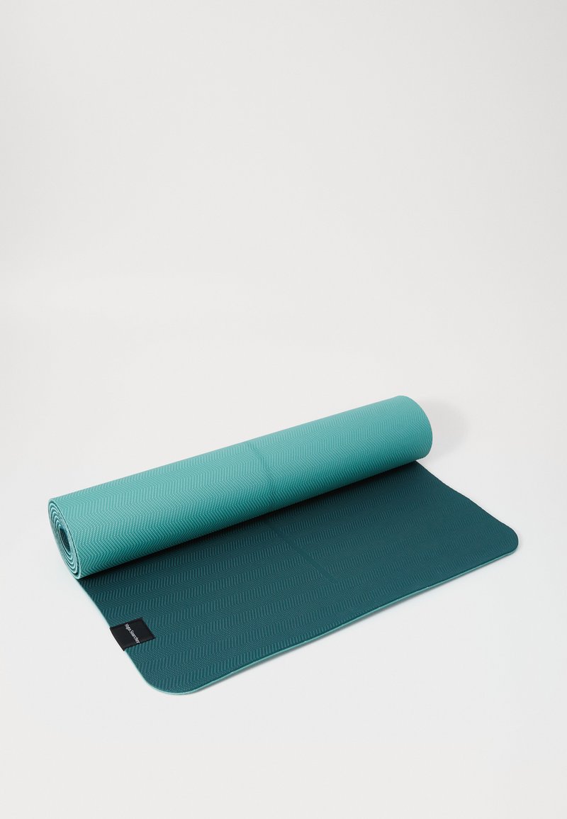 Esterilla de yoga con una superficie texturizada en color azul verdoso y verde oscuro, enrollada parcialmente, con una etiqueta visible cerca del borde.