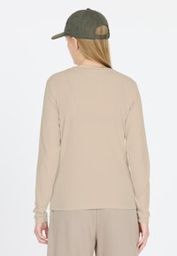 Beige, langärmliges, geripptes Oberteil mit rundem Ausschnitt, getragen zu grauen Hosen. Eine dunkelgrüne Mütze rundet das Outfit ab, von hinten betrachtet.