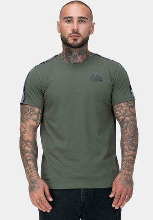 Bawełniana koszulka w kolorze oliwkowej zieleni z krótkimi rękawami, z bocznymi paskami i logo na piersi, noszona z czarnymi dżinsami.