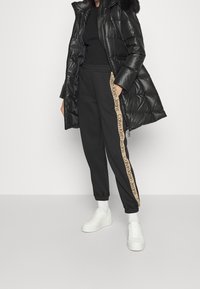 Nafoukaný černý kabát s kapucí lemovanou kožešinou, doplněný o černé tepláky s béžovým proužkem s logem a bílé tenisky.